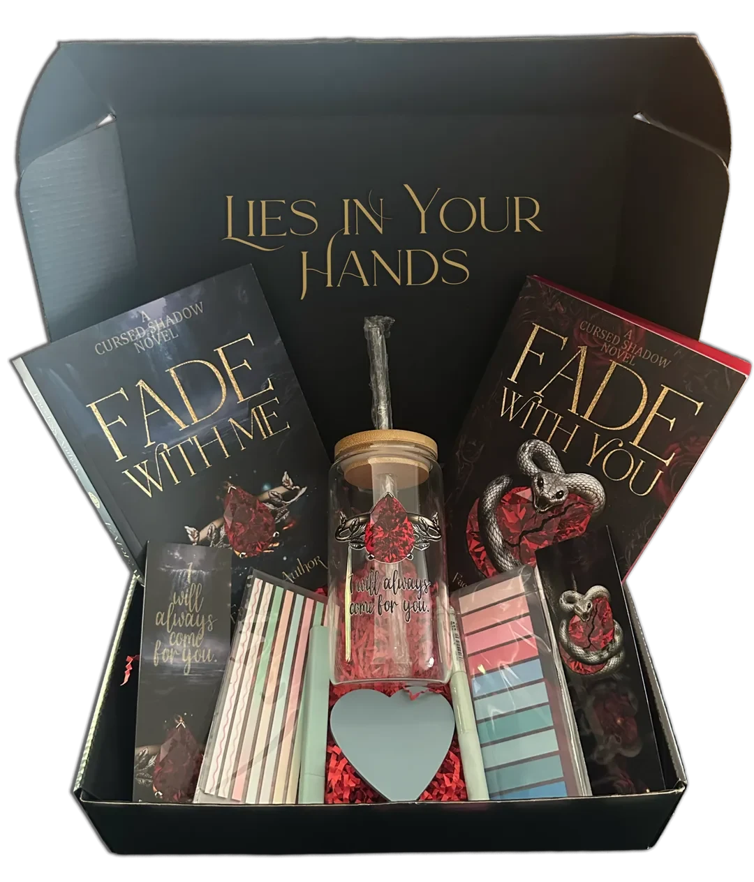 The Cursed Shadow Collector’s Box, open. The lid reads “Lies in Your Hands”. Inside: Fade With Me and Fade With You, a glass cup with a bamboo lid and a red gem design, two bookmarks, page flags, sticky tabs and a heart-shaped note pad on red paper shred.