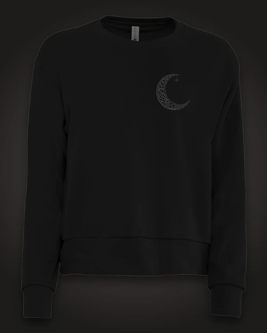 Mockup, front: black women’s crewneck with a small ornate crescent moon on the left chest.